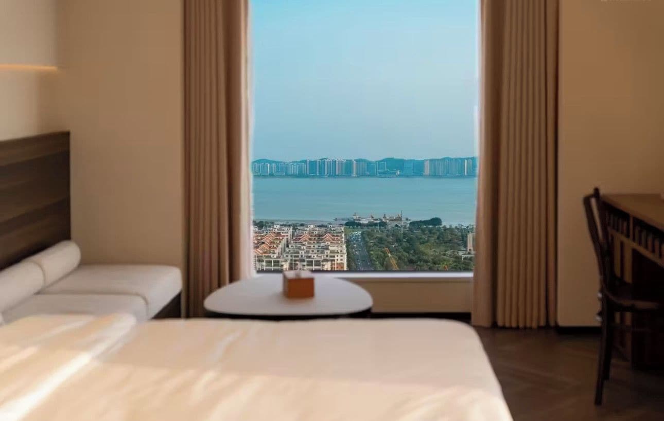 Quarto de hotel com cama, mesa redonda pequena, uma cadeira e uma janela grande com vista para a paisagem urbana e uma ponte.