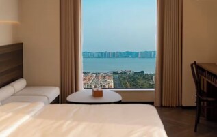 A hotel room with a bed, a small round table, a chair, and a large window offering a view of a cityscape and a bridge.