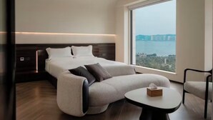 Quarto de hotel moderno com uma cama grande, um sofá, uma mesinha e vista para a cidade através de uma janela ampla.