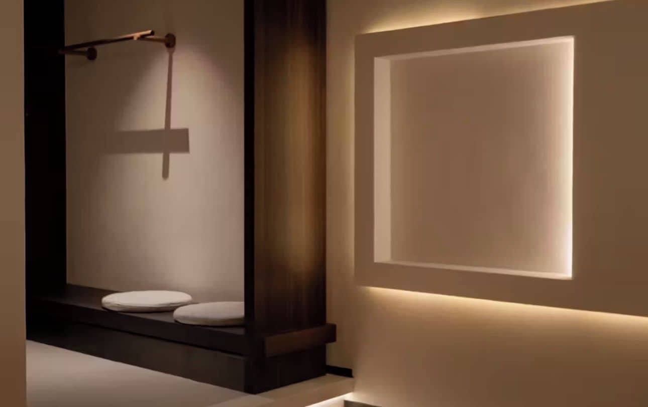 A modern bathroom with a minimalist design, featuring a wooden bench, a towel rack, and a wall-mounted light fixture.