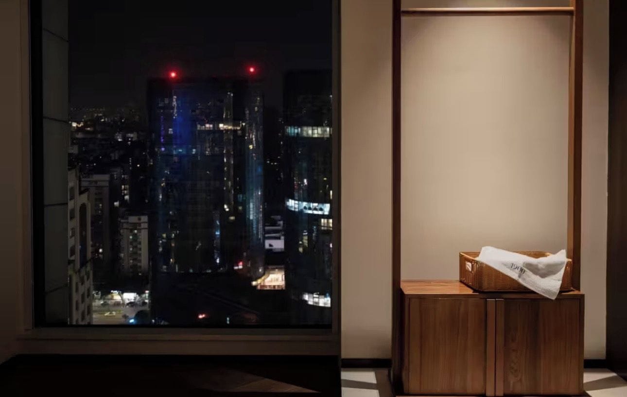 Ein Zimmer mit einem großen Fenster mit Blick auf eine nächtliche Stadtlandschaft, einem Holfnachtkästchen mit Taschentüchern und einer schlichten Wand.