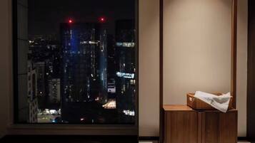 Ein Zimmer mit einem großen Fenster mit Blick auf eine nächtliche Stadtlandschaft, einem Holfnachtkästchen mit Taschentüchern und einer schlichten Wand.