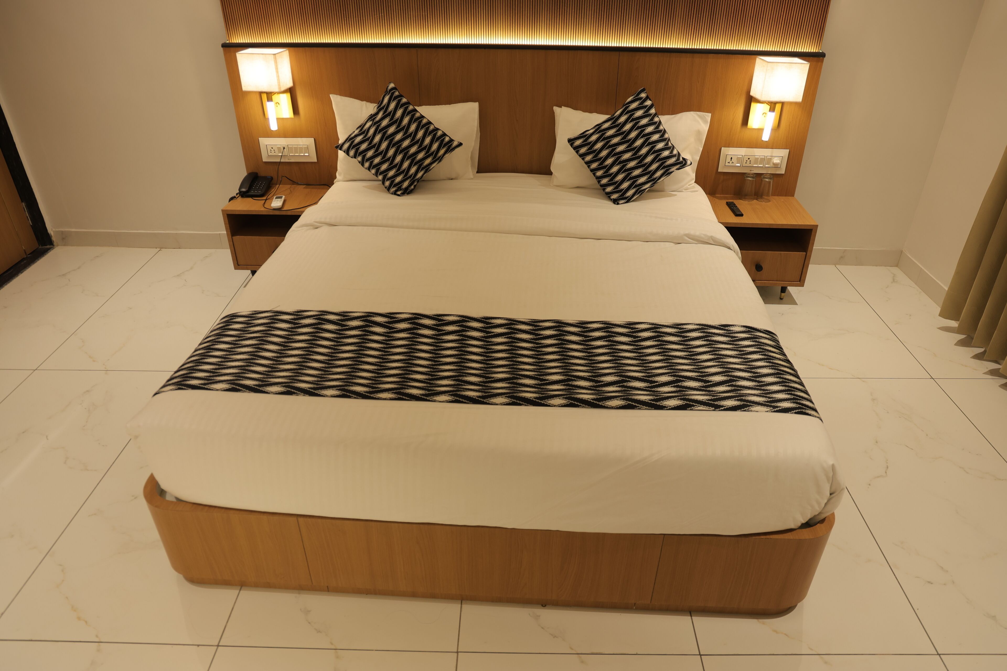 A hotel room with a large bed, two bedside tables, a wooden headboard, and a patterned throw blanket.