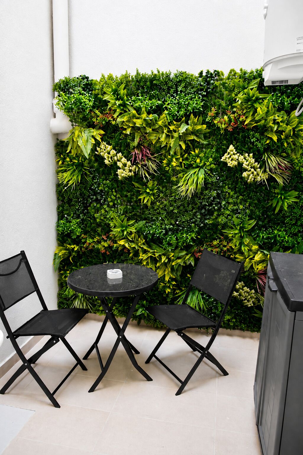Un pequeño espacio al aire libre con una mesa redonda y dos sillas, un muro vertical cubierto de plantas verdes y una pared blanca con una lámpara.