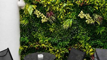 Un pequeño espacio al aire libre con una mesa redonda y dos sillas, un muro vertical cubierto de plantas verdes y una pared blanca con una lámpara.