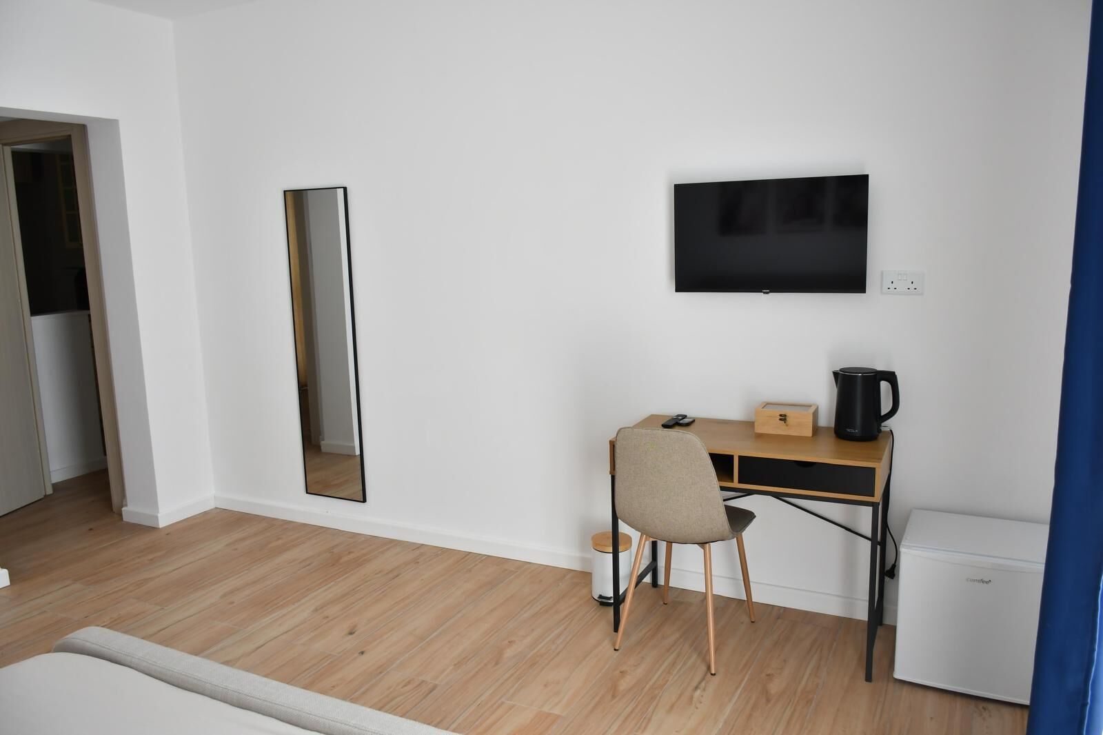 A room with a wooden desk, a chair, a flat-screen TV mounted on the wall, a small white appliance, and a mirror.