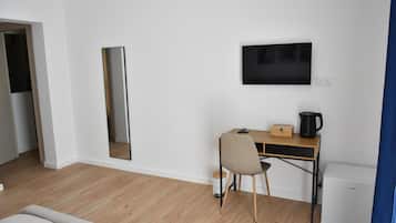 A room with a wooden desk, a chair, a flat-screen TV mounted on the wall, a small white appliance, and a mirror.