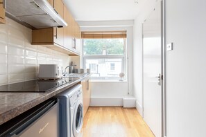 A modern kitchen with a built-in oven, a washing machine, and a window with a view of a house. - Cosy studio Apartment near Hyde Park (London)