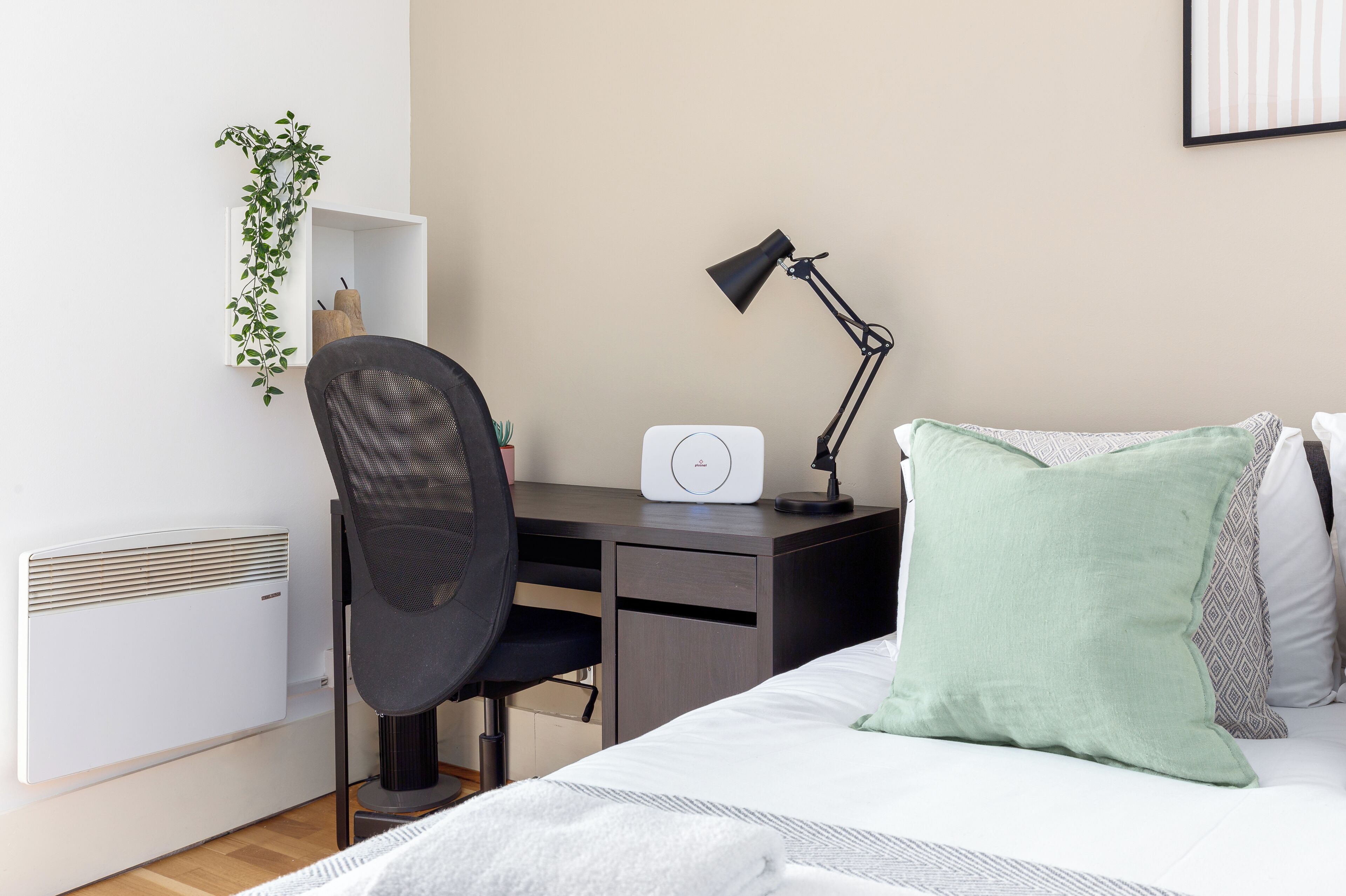 A bedroom with a bed, a desk, a chair, a lamp, a radiator, and a wall-mounted shelf with a plant.