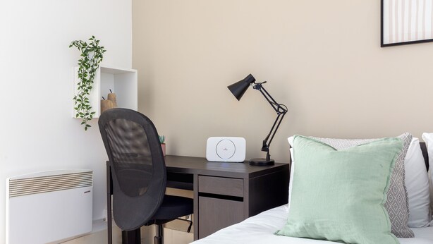 Quarto com cama, escrivaninha, cadeira, abajur, radiador e prateleira na parede com uma planta.