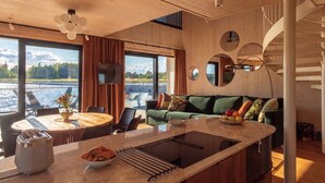 A modern interior with a wooden ceiling, a dining area with a round table, a sofa with cushions, and a view of a body of water through large windows. - Stay North - Loviisa Floating House (Loviisa)