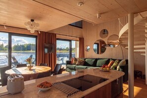 A modern interior with a wooden ceiling, a dining area with a round table, a sofa with cushions, and a view of a body of water through large windows. - Stay North - Loviisa Floating House (Loviisa)