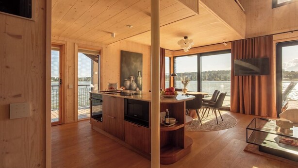 A modern interior with a wooden ceiling, a dining area with a view of the water, and a kitchen with wooden cabinets and a large window. - Stay North - Loviisa Floating House (Loviisa)