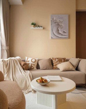 A cozy living room with a sofa, a round white table with a bowl of fruit, a bookshelf with plants, and a wall-mounted artwork. - 3BR/4Beds | 2BA • Central D1 • Group Stay • Corner (Thành phố Hồ Chí Minh)