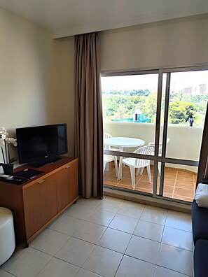 Ein Hotelzimmer mit Fernseher auf einer Holzkommode, einem Balkon mit Tisch und Stühlen sowie Blick auf Grünflächen und Gebäude.