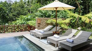 Un área junto a la piscina con sillones de descanso y una sombrilla, rodeada de exuberante vegetación.