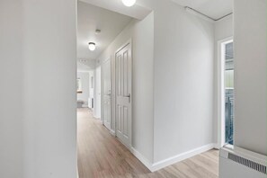 Ein Korridor mit weißen Wänden und Holzdielen, der zu einem Zimmer mit Fenster und Heizkörper führt.