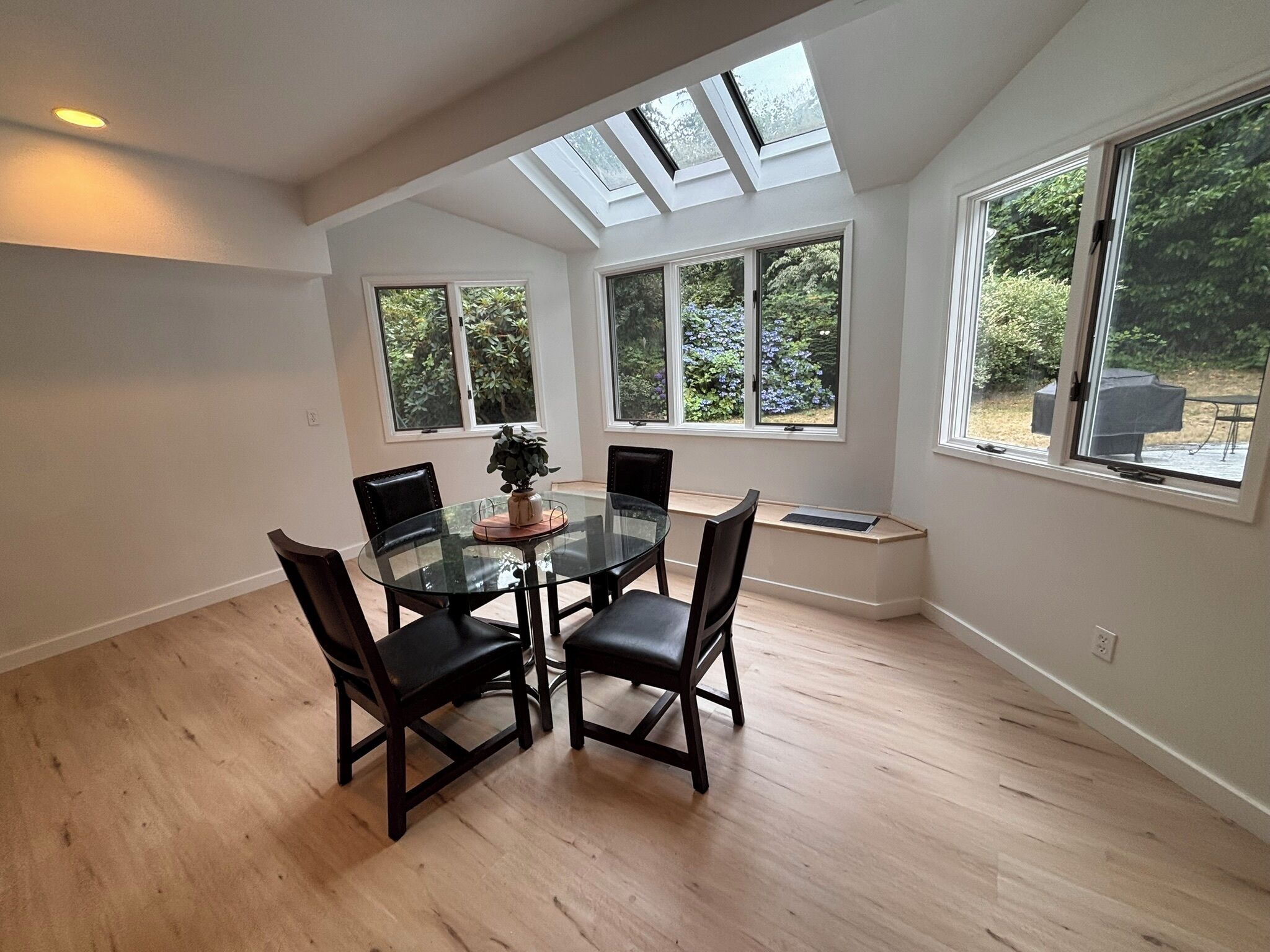 A dining area with a glass table and chairs, large windows with views of greenery, and a skylight.
