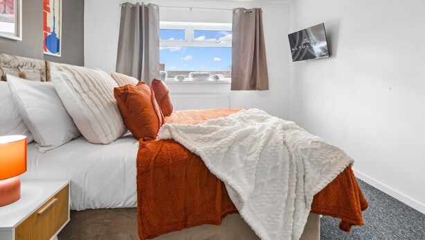 Habitación de hotel con cama, mesita de noche, lámpara, ventana con cortinas y televisor montado en la pared.