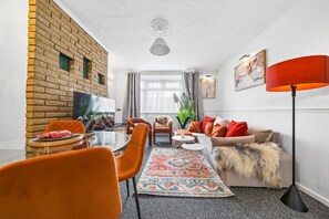 Ein modernes Wohnzimmer mit Ziegelwand, Flachbildfernseher, Esstisch mit Stühlen, Sofa mit bunten Kissen und Stehlampe.