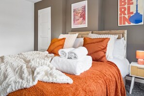 Ein ordentlich bezogenes Bett mit weißer und orangefarbener Bettwäsche, eine weiße Tür und ein gerahmtes Stadtbild an der Wand.