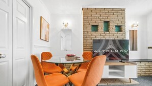 Un comedor con una mesa redonda de vidrio, sillas tapizadas en naranja, una pared de ladrillo decorativa y un televisor de pantalla plana.