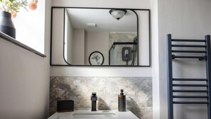 A bathroom with a large mirror, a towel rack, a sink with two soap dispensers, and a decorative vase with flowers. - Premium 1BD in Beaufort Square | Central Chestow (Monmouthshire)