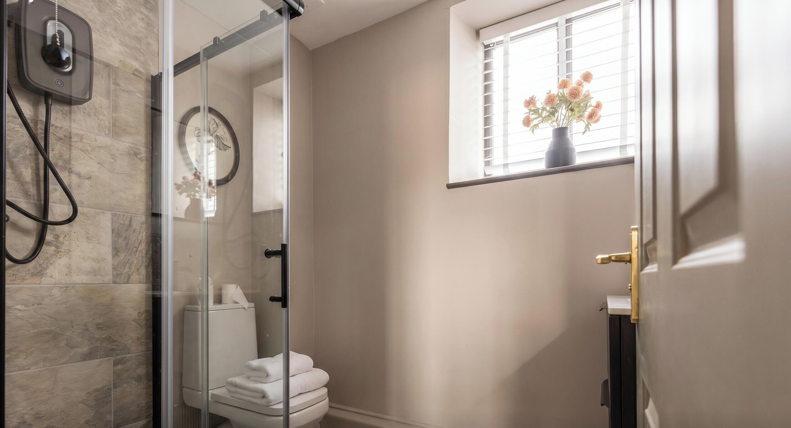 Ein modernes Badezimmer mit Duschkabine aus Glas, Toilette und einem Fenster mit Blumenvase.