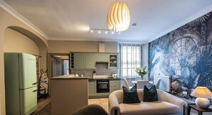 A modern kitchen with a central island, a dining area with a sofa and cushions, and a large tropical mural on the wall. - Premium 1BD in Beaufort Square | Central Chestow (Monmouthshire)