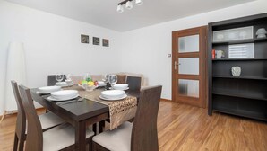 A dining area with a wooden table set for four, a sofa, and a bookshelf with framed items. - Spacious Apartment with Balcony | 3 Bedrooms | Workspace (Zabrze)
