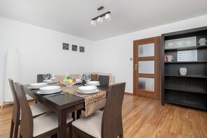 Un coin repas avec une table en bois dressée pour quatre personnes, un canapé et une bibliothèque agrémentée d’objets décoratifs encadrés.
