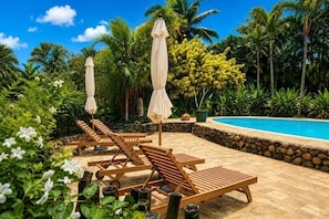 Zona junto a la piscina con sillones de descanso y sombrillas, rodeada de vegetación tropical y un muro de piedra.