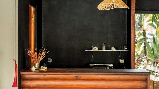 Een houten eettafel met stoelen, een hangende geweven lamp en een zwarte achtergrond met planken.