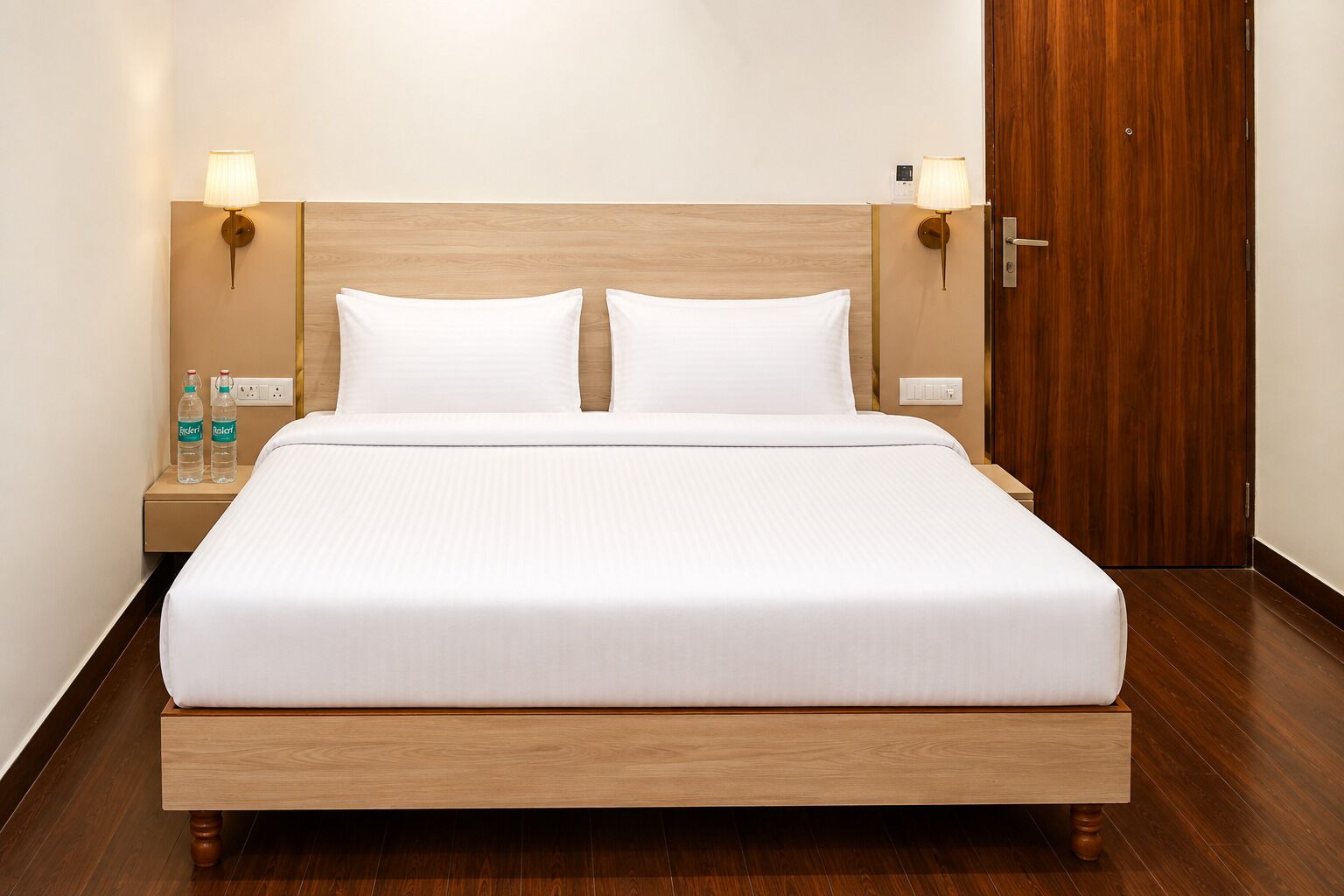 Una habitación de hotel con una cama grande, cabecera de madera, lámparas de noche, una mesita de noche con botellas de agua y una puerta de madera.