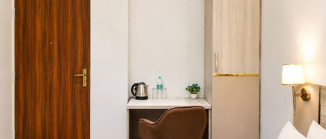 A hotel room with a wooden door, a desk with a chair, a small table with a kettle, a bottle, and a plant, a wardrobe, and a bedside lamp.