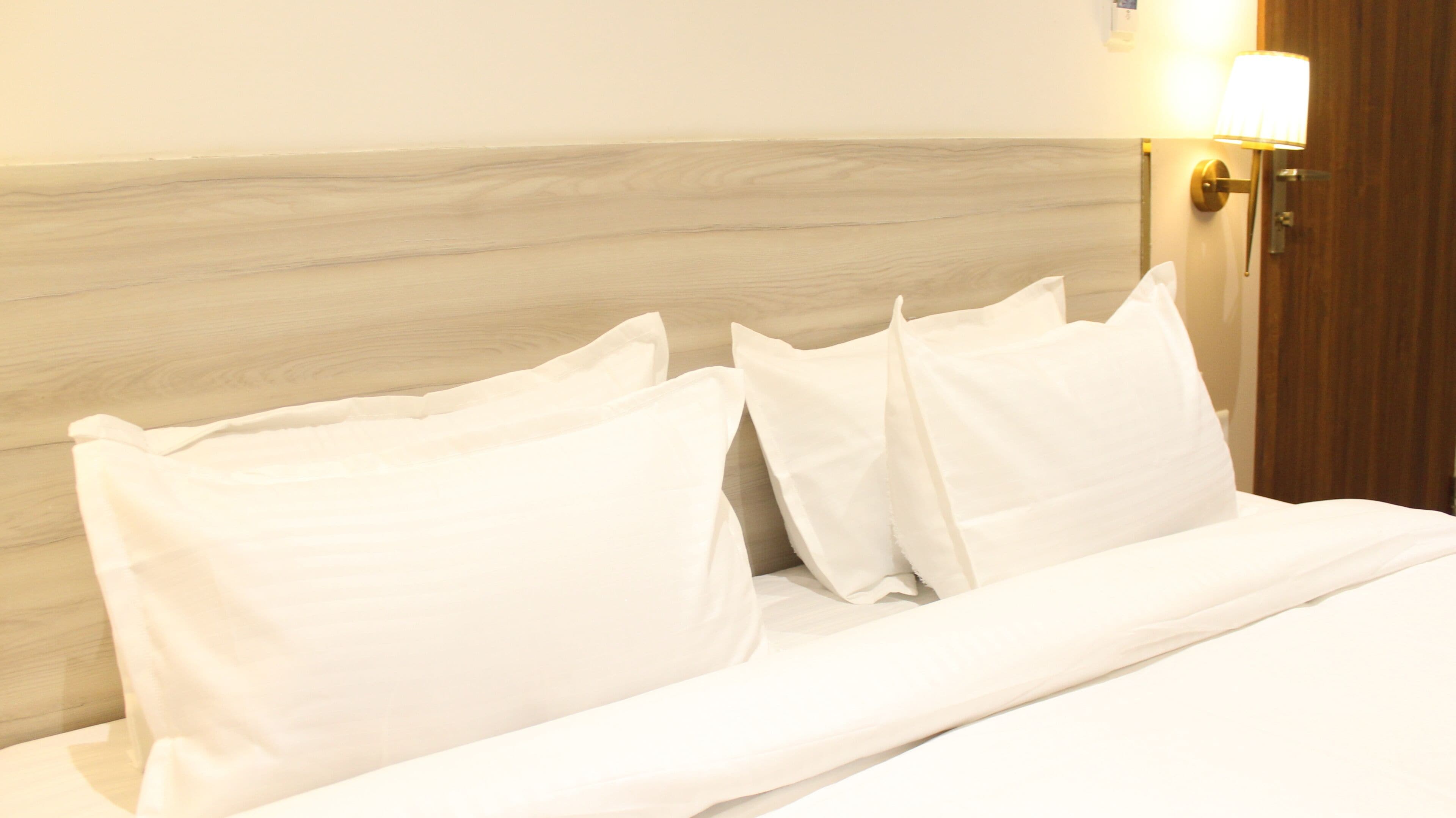 A neatly made bed with white linens and pillows, a wooden headboard, and a wall-mounted light fixture.