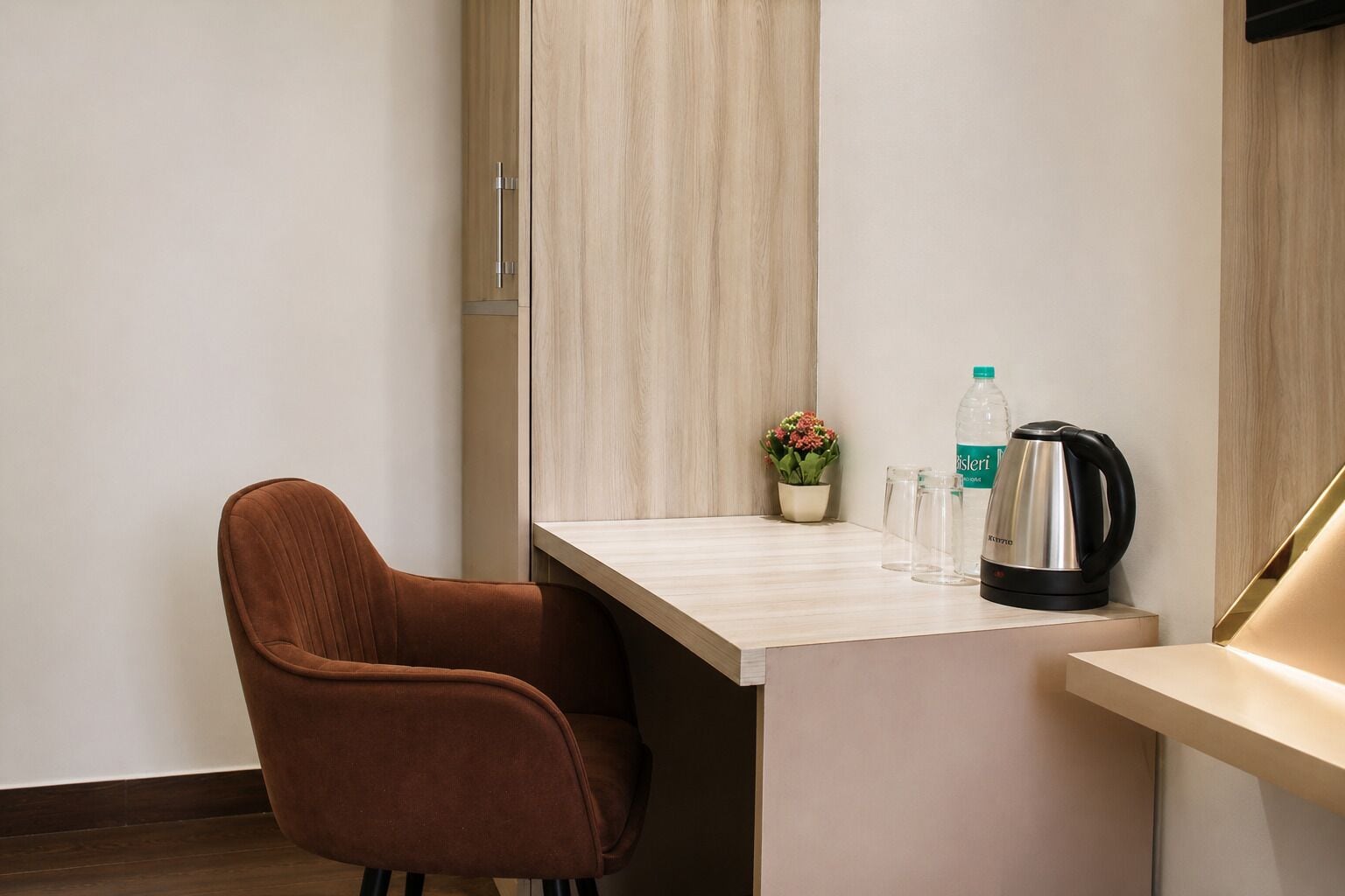 A hotel room with a brown upholstered chair, a wooden desk, a small potted plant, a water bottle, a glass, and a kettle.
