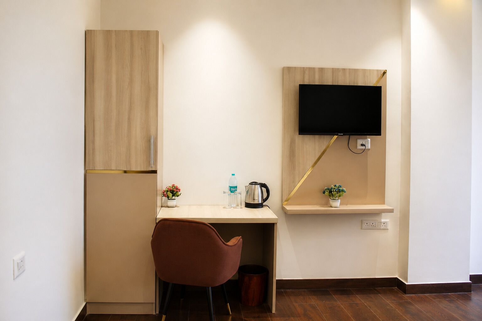 Una habitación pequeña y moderna con un armario de madera, un escritorio con una silla, un televisor montado en la pared y un estante con una planta.