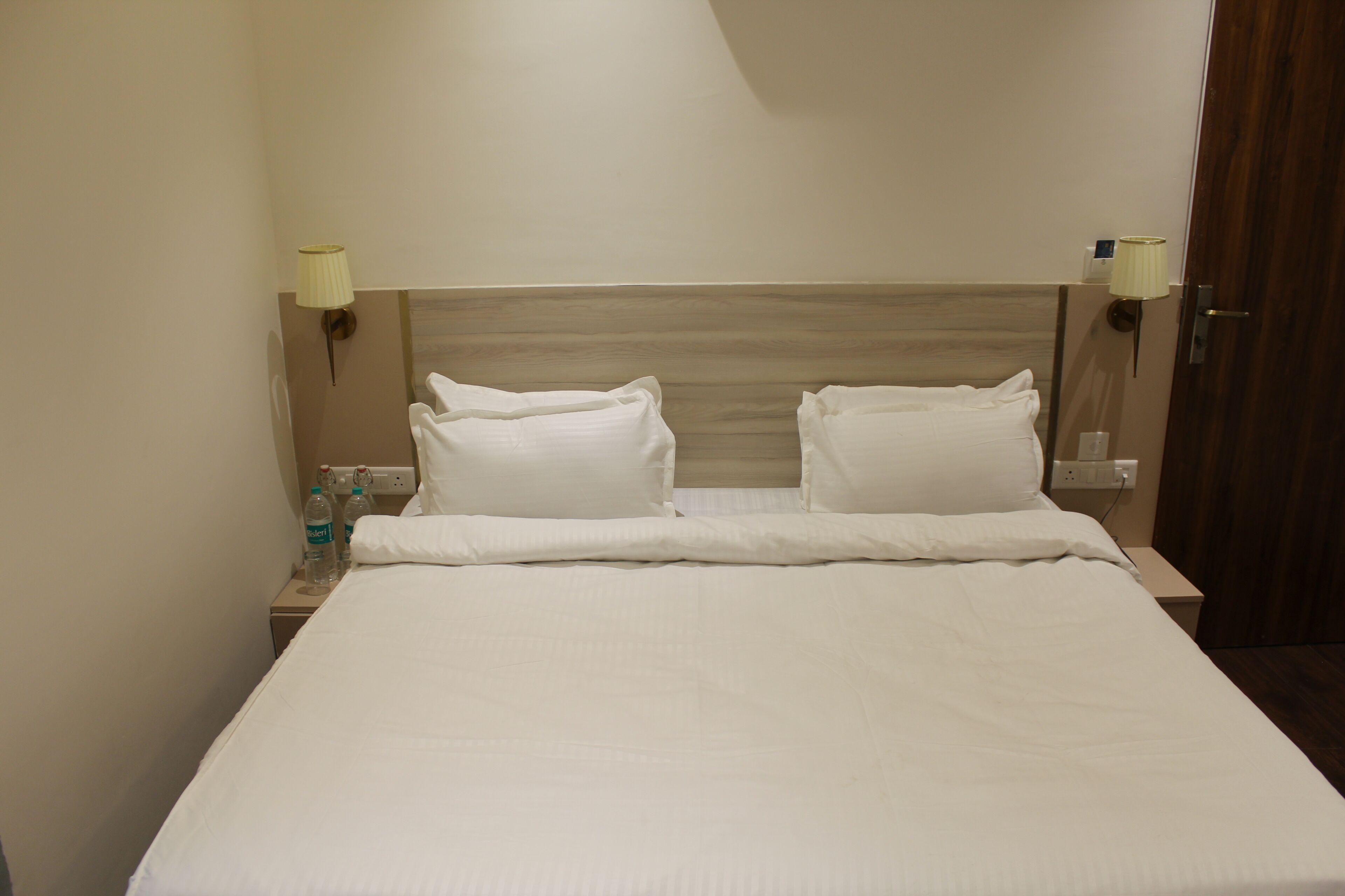 A hotel room with a bed, two bedside lamps, a nightstand with water bottles, and a wooden headboard.
