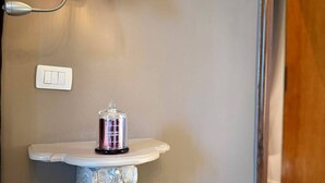 Un tocador de baño con un pedestal decorativo, una pequeña repisa con una botella, una lámpara de pared y un enchufe.