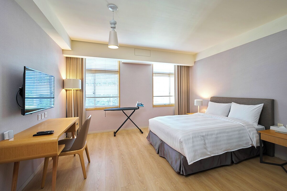 A hotel room with a bed, desk, chair, television, and a window with blinds.