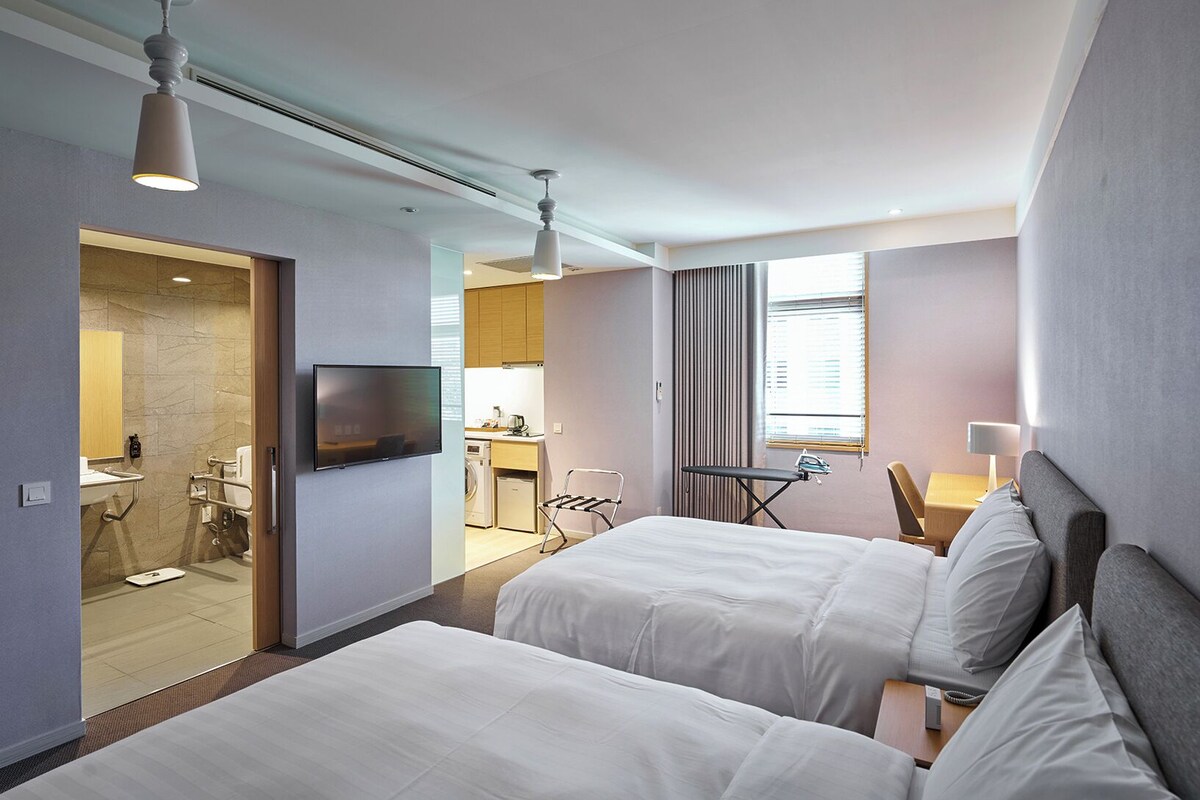 A hotel room with two beds, a TV, a desk with a chair, and a bathroom visible through an open door.
