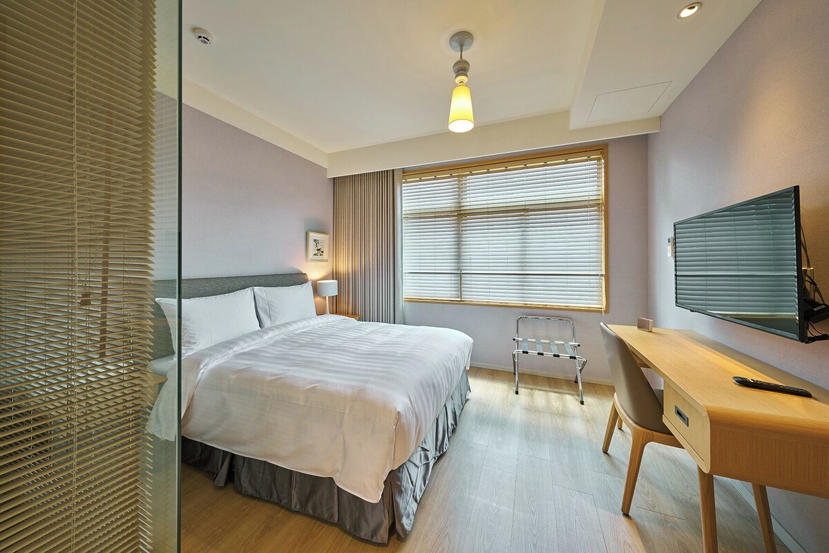 A hotel room with a bed, a desk, a chair, a television, and a window with blinds.