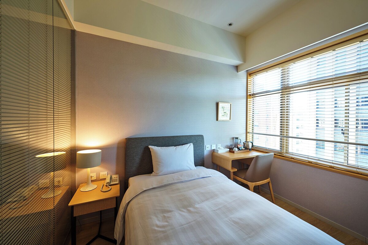 A hotel room with a bed, bedside table, lamp, and a window with blinds.