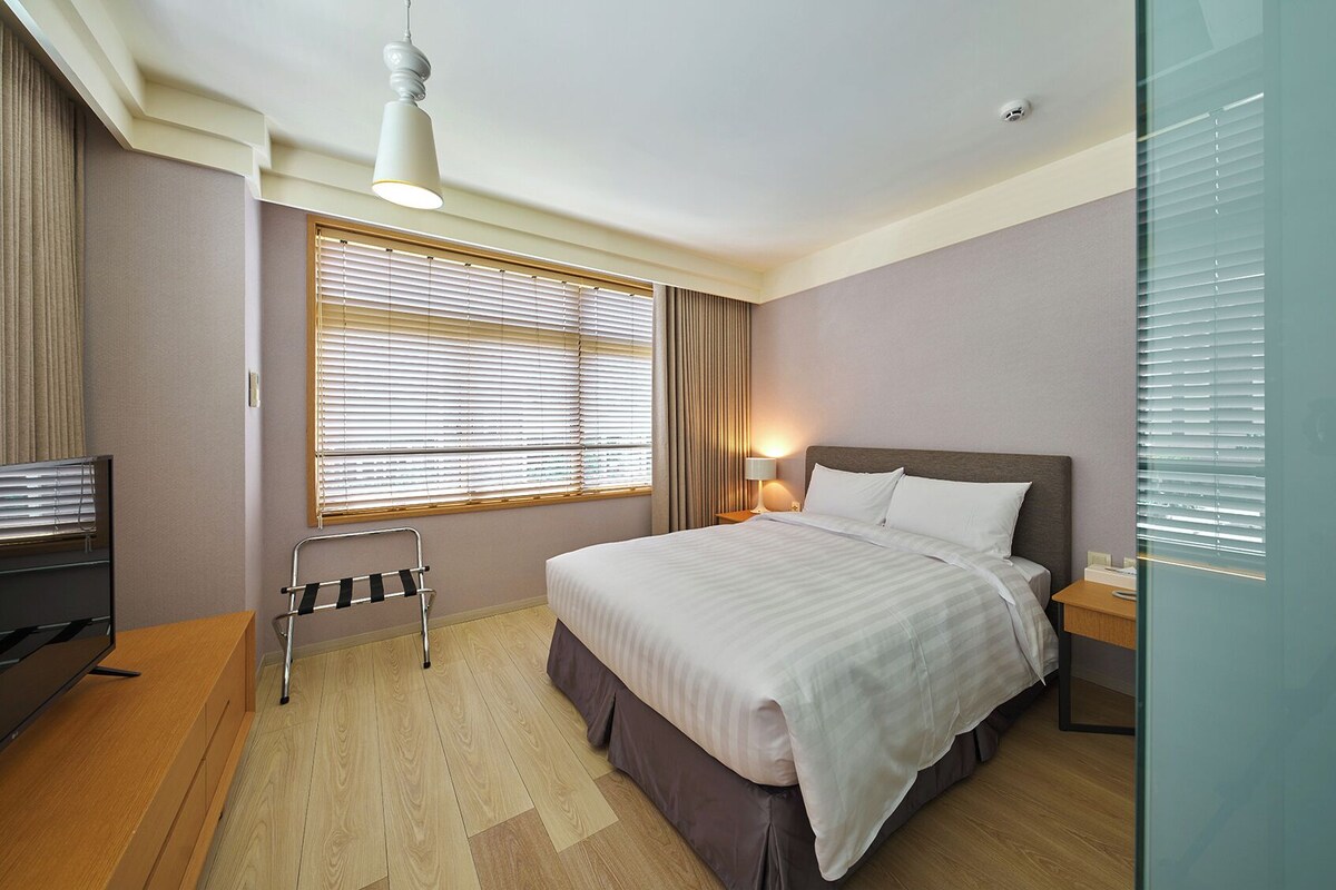 A hotel room with a bed, bedside table, lamp, TV, and a window with blinds.
