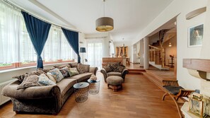 A spacious living room with a large sofa, a smaller armchair, and a coffee table. A staircase leads to an upper level, and a piano is visible in the background. - Apartamenty Sun & Snow Villa Łokietka (Sopot)