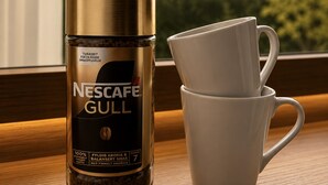 En Nescafe Gull kaffemaskine og to hvide kopper på en træoverflade ved et vindue med udsigt til træer.