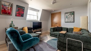 Un salon moderne avec un grand téléviseur à écran plat, un fauteuil bleu, un canapé vert et une affiche du « Club des amateurs de vin et de fromage » accrochée au mur.