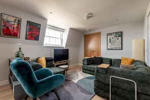 A modern living room with a large flat-screen TV, a blue armchair, a green sofa set, and a 'Wine & Cheese Club' poster on the wall. - Century Court (No 10) | St Andrews (St Andrews)