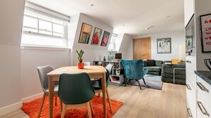 Un appartement moderne comprenant un coin repas, un salon et une cuisine. Les murs sont ornés de tableaux encadrés, et on y trouve une table à manger en bois avec des chaises, ainsi qu’un canapé dans le salon.